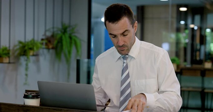 Caucasian businessman reviews printed documents by laptop in modern coworking office, turning pages and comparing work materials for project planning, report analysis and professional productivity.