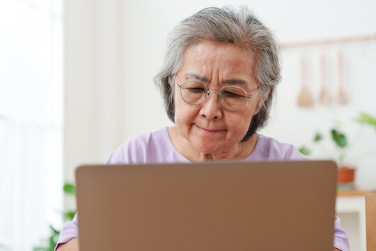 Close up of senior Asian woman with gray hair and eyeglasses working, looking at computer laptop in bright cozy, modern living room with focus and concentration