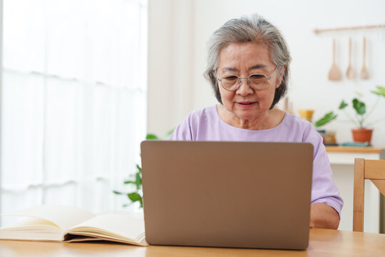 Senior Asian woman with gray hair and eyeglasses sitting, working and typing on computer laptop on wooden table in bright cozy, modern living room with joy. Active lifestyle and technology