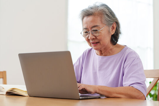 Senior Asian woman with gray hair and eyeglasses sitting, working and typing on computer laptop on wooden table in bright cozy, modern living room with joy. Active lifestyle and technology