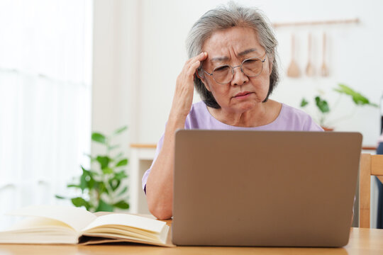 Stressed, tired senior Asian woman with gray hair and eyeglasses working, looking at computer laptop, put her hand on forehead in confusion in bright cozy, modern living room