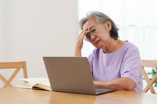 Stressed, tired senior Asian woman with gray hair and eyeglasses working, looking at computer laptop, put her hand on forehead in confusion in bright cozy, modern living room