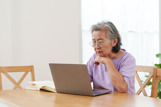 Senior Asian woman with gray hair and eyeglasses sitting at wooden table, learning, watching online learning course on computer laptop in bright cozy, modern living room with focus and concentration