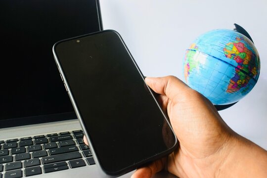 Hand holding a black smartphone with a laptop and a desktop globe in the background, representing global connectivity, mobile work, and digital communication.