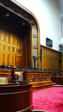 Main chamber of Belgrade's National Assembly: speaker's podium and rows of delegate seats with screens and microphones. Depicts legislative proceedings, civic institutions, and democratic governance.