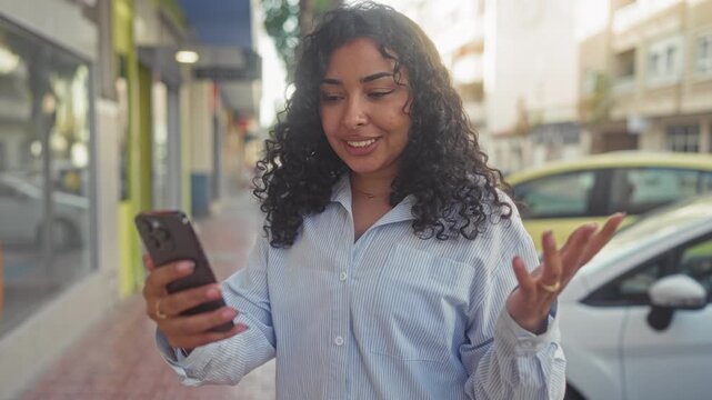 Young arab woman holding smartphone and scrolling, gesturing while checking messages on a busy street lined with storefronts and parked cars; casual connection.