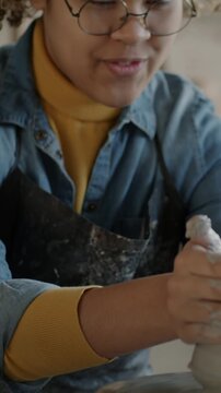 Tilt-down portrait of joyful young lady in apron working with clay making pot on throwing wheel in workshop. Creative person and hobby concept.