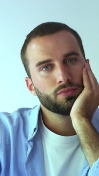 Pensive young bearded man with blue eyes looking at the camera and then away, expressing boredom and thoughtfulness while resting his head on his hand against a clean white background