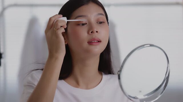 Young Asian woman looking into a vanity mirror while applying mascara or lash serum to her eyelashes.