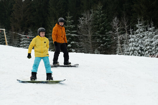 Two snowboarders glide down a snowy slope surrounded by evergreen trees, enjoying a winter adventure on a sunlit day in the mountains