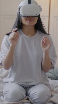 Slow motion of girl in pajamas sitting on bed using virtual reality glasses enjoying new experience in bedroom at home. Modern technology and entertainment concept.