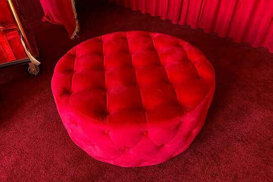A round red velvet tufted ottoman sits in a room with a red carpet floor and red background