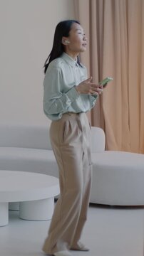 Cheerful Asian lady talking on mobile phone using wireless earphones and smartphone walking in luxury apartment. Communication and people concept.