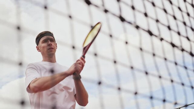 Young adult Caucasian man hitting tennis ball with racket on court in slow motion, low angle view through net. Footage suited for sports marketing and active lifestyle fitness campaigns
