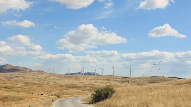 Wind Turbines on Dry Hills Under Blue Sky. Wind turbines standing across dry rolling hills under a blue sky with clouds, with a country road leading through the rural landscape.