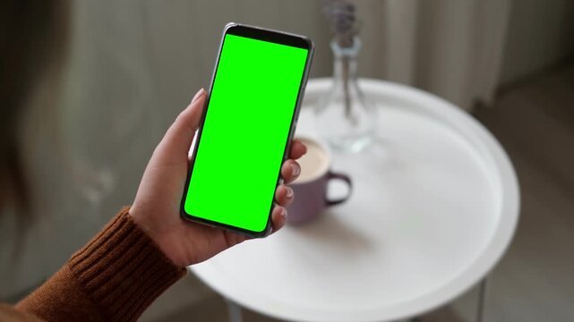 Top view of woman holding smartphone with vertical green screen chroma key inside cozy room. Girl sitting on sofa using mobile phone template mockup display next to a coffee cup on white round table