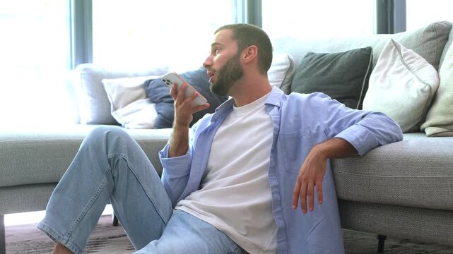 Handsome casual man sitting on the floor in his living room, leaning against the couch and talking on the speakerphone, sending a voice message or having a relaxed conversation at home