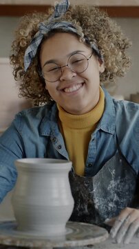 Slow motion portrait of happy woman sitting at work table in pottery workshop smiling looking at camera. African American female potter smiling enjoying creative activity.