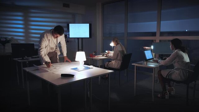 An electronics specialist reviews a control electronics scheme at a large table while two colleagues work on laptops in a nighttime laboratory setting with large windows.