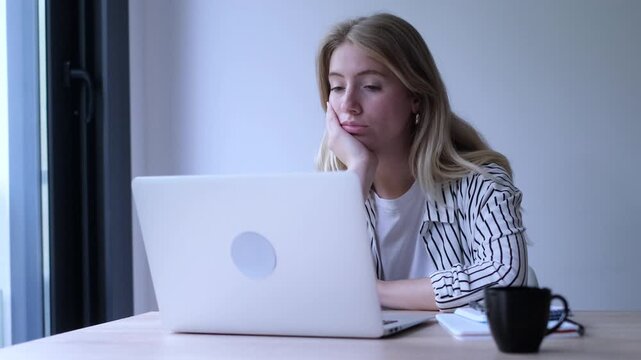 Frustrated young female employee feeling bored and unhappy while working on a laptop computer, experiencing burnout and procrastination from her unfulfilling office job at the table