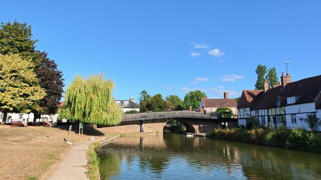 Hungerford Berkshire England UK. 28.08.2026. Video. A waterside view featuring a large weeping willow tree, traditional English houses, and a calm canal reflecting the blue sky and clouds