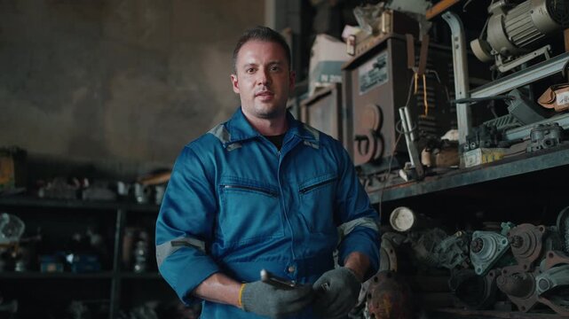Focused mechanic stands in cluttered workshop. Shelves hold tools and broken parts. Dark space shows worn machinery and grease