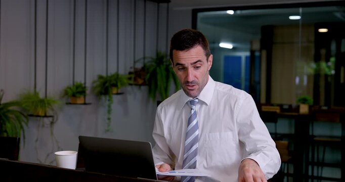 Caucasian businessman sorts printed charts beside laptop in modern coworking office, lifting report pages and comparing business data for document review, project planning and professional analysis.