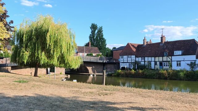 Hungerford Berkshire England UK. 28.08.2026. Video. A waterside view featuring a large weeping willow tree, traditional English houses, and a calm canal reflecting the blue sky and clouds