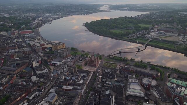 Aerial view of Peace Bridge spanning the River Foyle with historic city buildings and lush green banks in Derry, Northern Ireland, United Kingdom.
