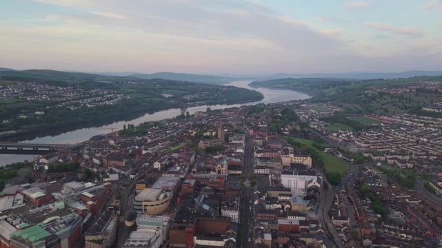 Aerial view of the city of Londonderry and River Foyle with historic buildings, bridges, and lush green hills under a soft sunset sky in Londonderry, United Kingdom.