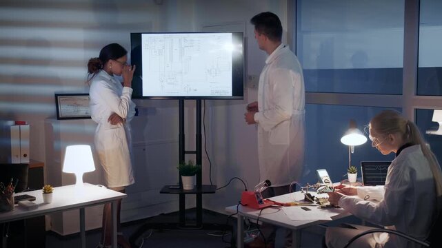 Three electronics engineers in white lab coats discuss complex technical schematics on a large monitor inside a bright, modern research laboratory during daytime.