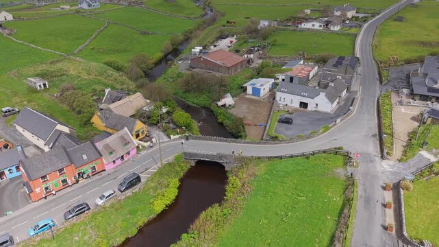 Aerial view of the Wild Atlantic Way passing through a village with a stone bridge over a river, colorful buildings, and lush green fields in Galway, County Galway, Ireland.
