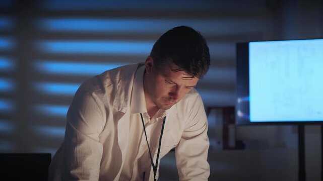 An engineer in a white shirt studies electronic schematics displayed on a large digital screen inside a professional workspace during nighttime.