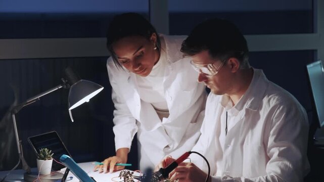Two electronics engineers collaborating on circuit boards and testing devices in a modern indoor laboratory setting during daytime.