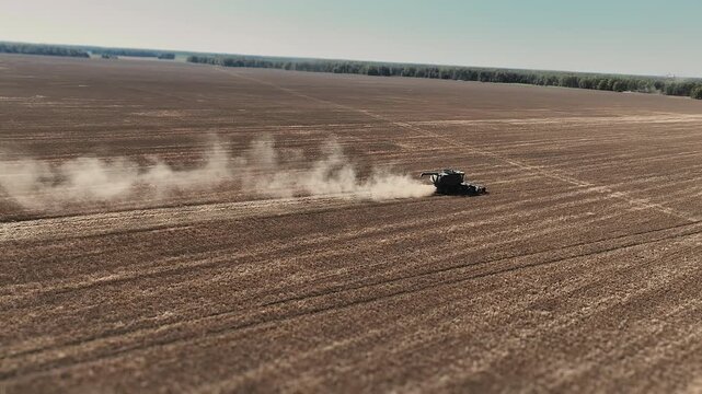 Aerial view of combine harvester harvesting flax crop across a large agricultural field, raising dust during seasonal farm work and crop production.