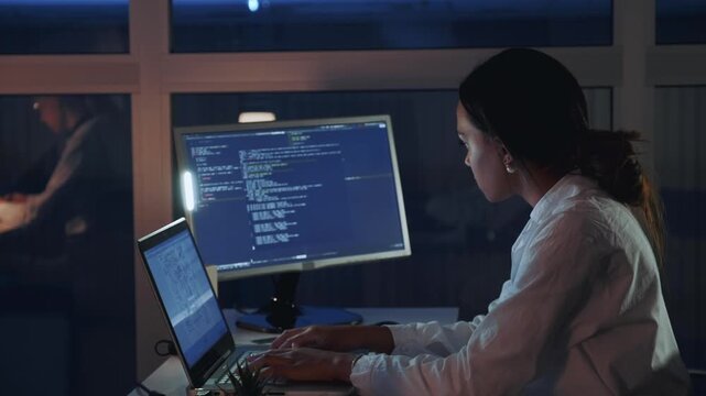 African American female engineer wearing lab coat coding on multiple monitors at desk in a nighttime electronics laboratory with large windows and a potted plant.