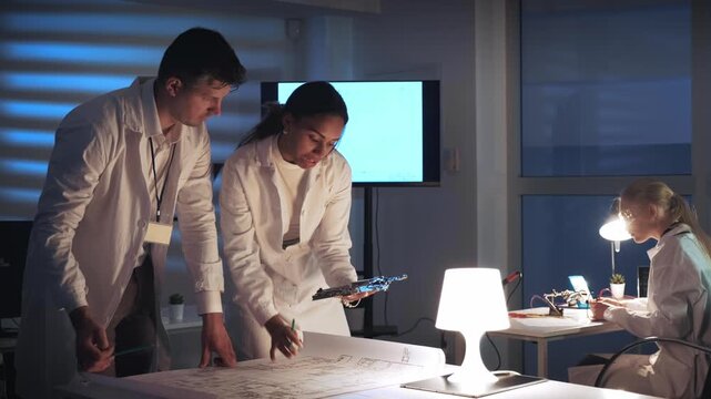 Three engineers in lab coats collaborate on electronics prototypes and technical schematics in a professional laboratory setting during daytime.