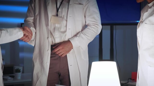 Scientists wearing white lab coats greet and interact inside a modern laboratory with technical equipment and illuminated lighting at nighttime.