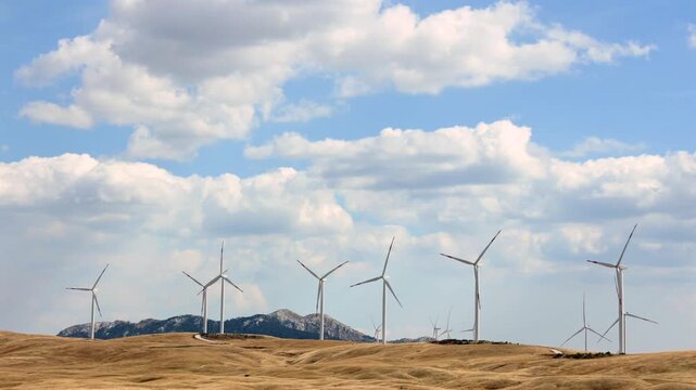 Wind Farm on Rolling Hills Under Cloudy Sky. Wind turbines standing on dry rolling hills with mountains and clouds in the background. Renewable energy and clean power landscape.