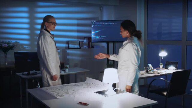 Two women in white lab coats collaborate on electronics schematics at a large table in a modern laboratory with technical equipment and natural daylight.