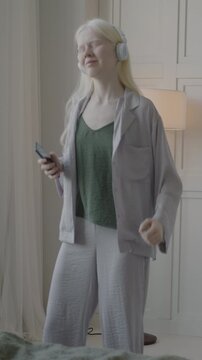 Beautiful albino lady wearing pajamas dancing to music in luxury bedroom using smartphone and headphones relaxing in weekend. Youth culture and people concept.