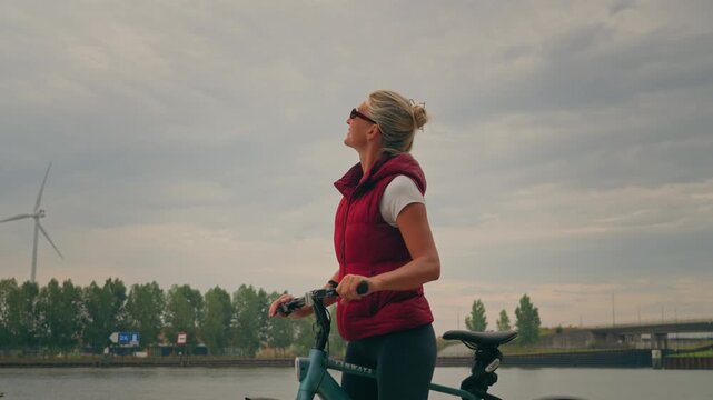 Caucasian blonde woman cycles into frame, brakes and dismounts before looking around and taking in the beautiful riverside views during a peaceful break.