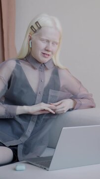 Cheerful albino girl making online video call with laptop and wireless earphones sitting on couch in luxury interior. Virtual meeting and accommodation concept.