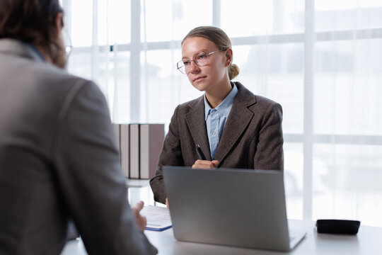 Young hr manager in professional attire listens and takes notes with pen and laptop while interviewing a male candidate across a desk in a focused hiring meeting setting