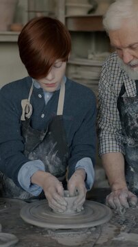 Kid making bowl from clay under guidance of caring grandpa in modern pottery studio. Boy making unique artwork using spinning wheel.
