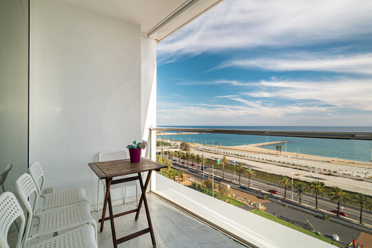 Modern balcony with white chairs and wooden table overlooking beach, ocean, blue sky, clouds, glass railing, potted plant, relaxing coastal resort setting, with balcony, chair, table, wooden detail