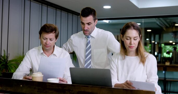 Three Caucasian business colleagues compare printed documents beside laptop in modern coworking office, pointing at paperwork and exchanging ideas during report review, planning and team discussion.