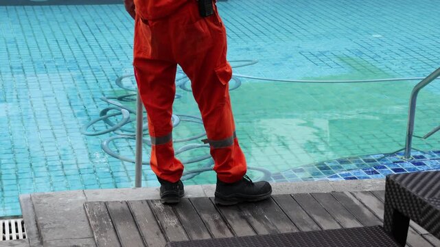 Worker in orange suit cleans swimming pool. He uses long pole to scrub pool surface. Pool water appears green and murky. Metal ladder sits beside tiled edge. Worker wears bright cap and sturdy boots