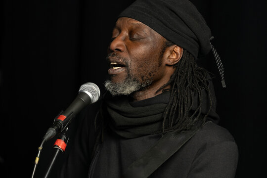 Close-up portrait of a black male singer performing live into a microphone. Emotional vocal performance in dark studio, minimal background, expressive face and authentic concert mood.