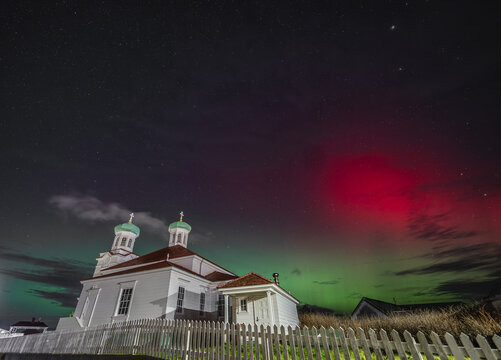 View of the historic Holy Ascension of Our Lord Cathedral illuminated by the vibrant green and red aurora borealis in the night sky, Unalaska, Alaska, United States.